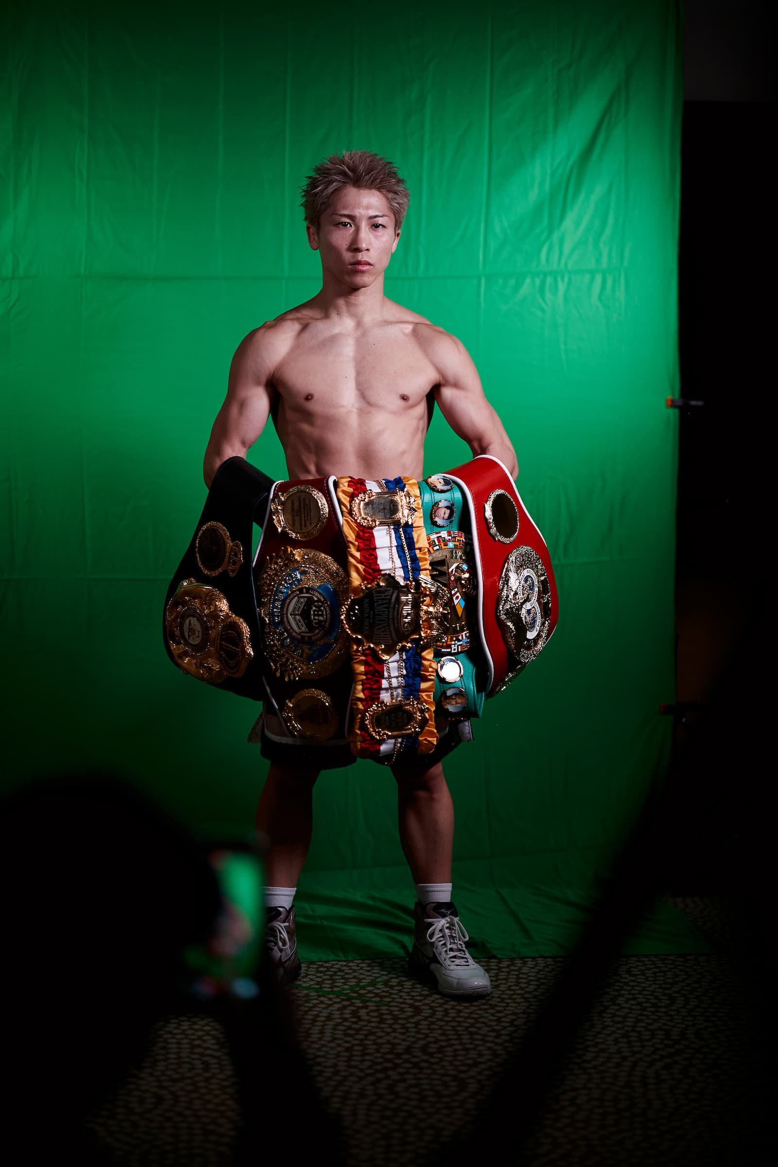 Naoya Inoue posing in front of a green screen with his championship belts