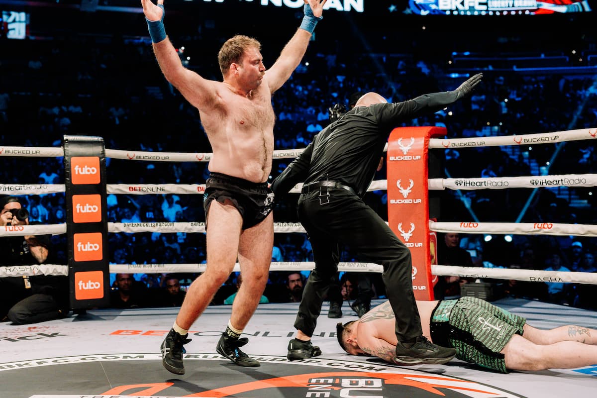 Lex Ludlow celebrates victory with his hands up while Drew Nolan is on the ground attended by the referee